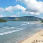 Vista panorâmica da Praia das Toninhas em Ubatuba com mar calmo, faixa de areia clara e famílias aproveitando o dia de sol, ideal para crianças.